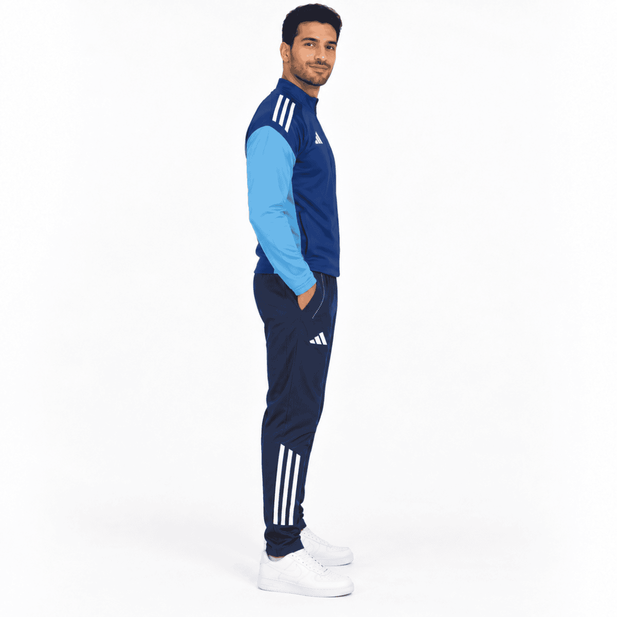 Man wearing blue Adidas tracksuit with white stripes, white sneakers, standing sideways.