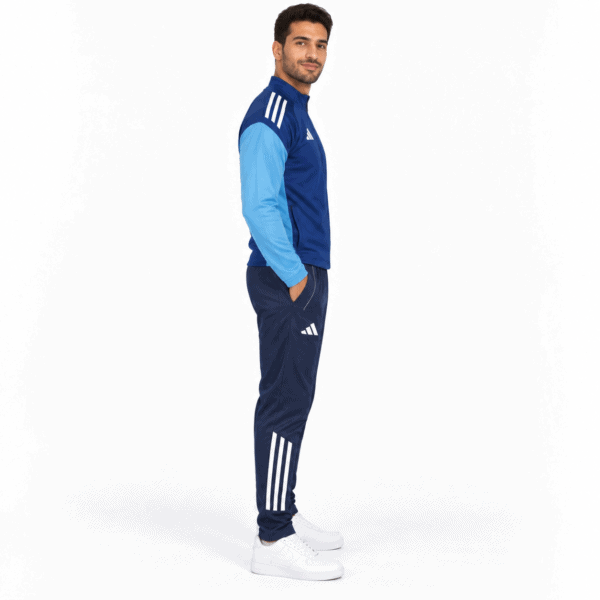Man wearing blue Adidas tracksuit with white stripes, white sneakers, standing sideways.