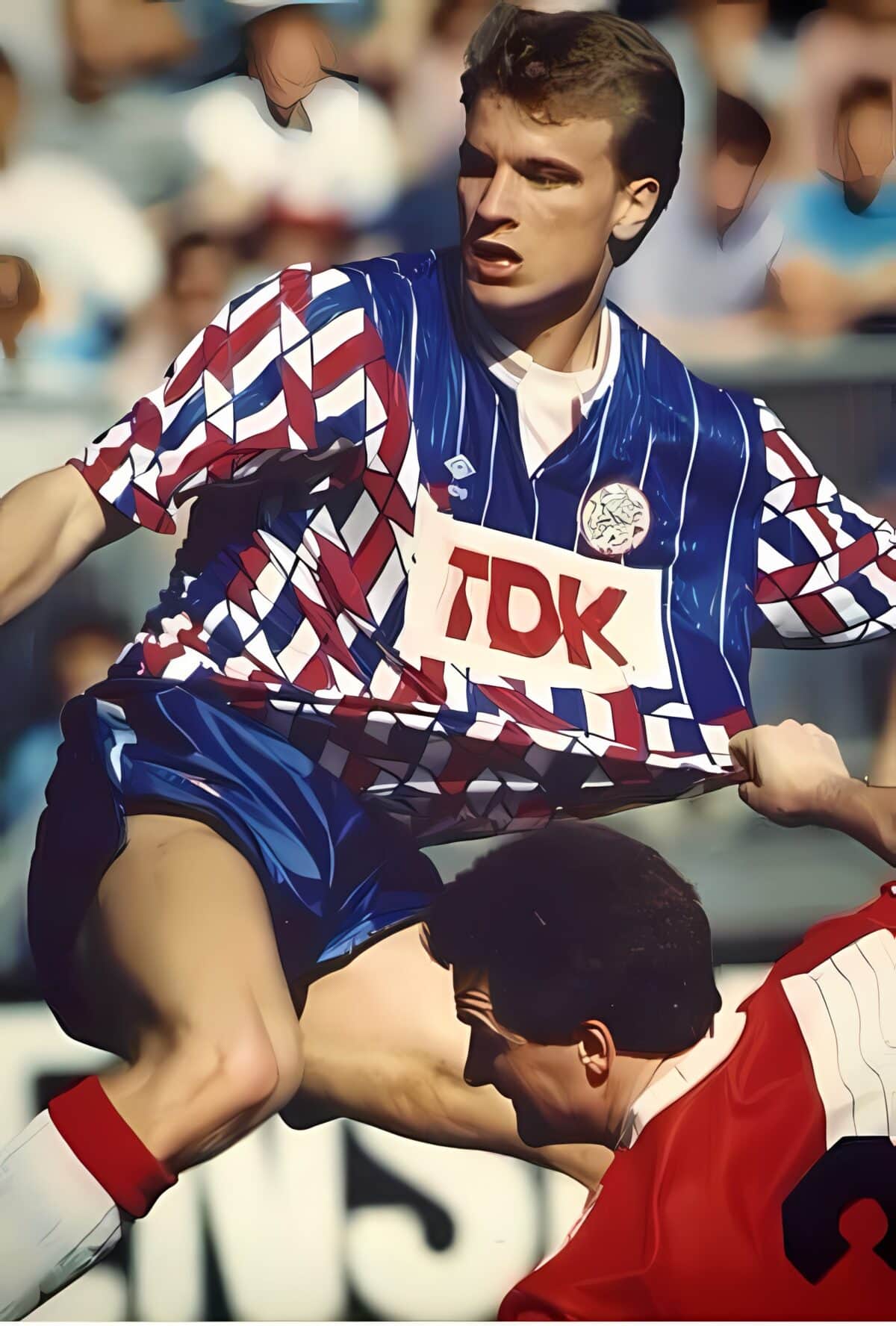Football player in blue and red kit during match with crowd in background.