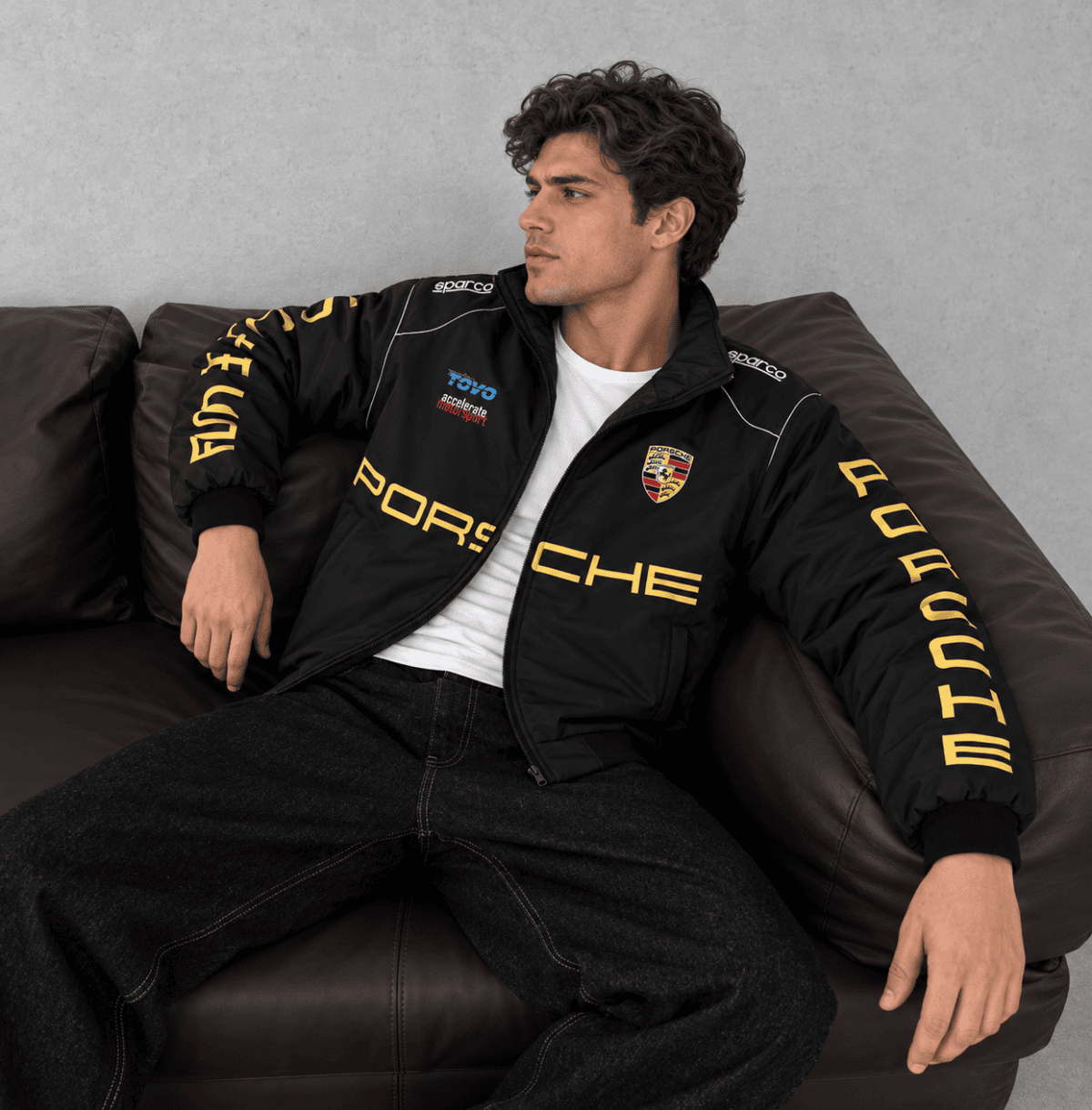 Man wearing Porsche Motorsport black jacket with yellow logo and crest, sitting on a brown leather.