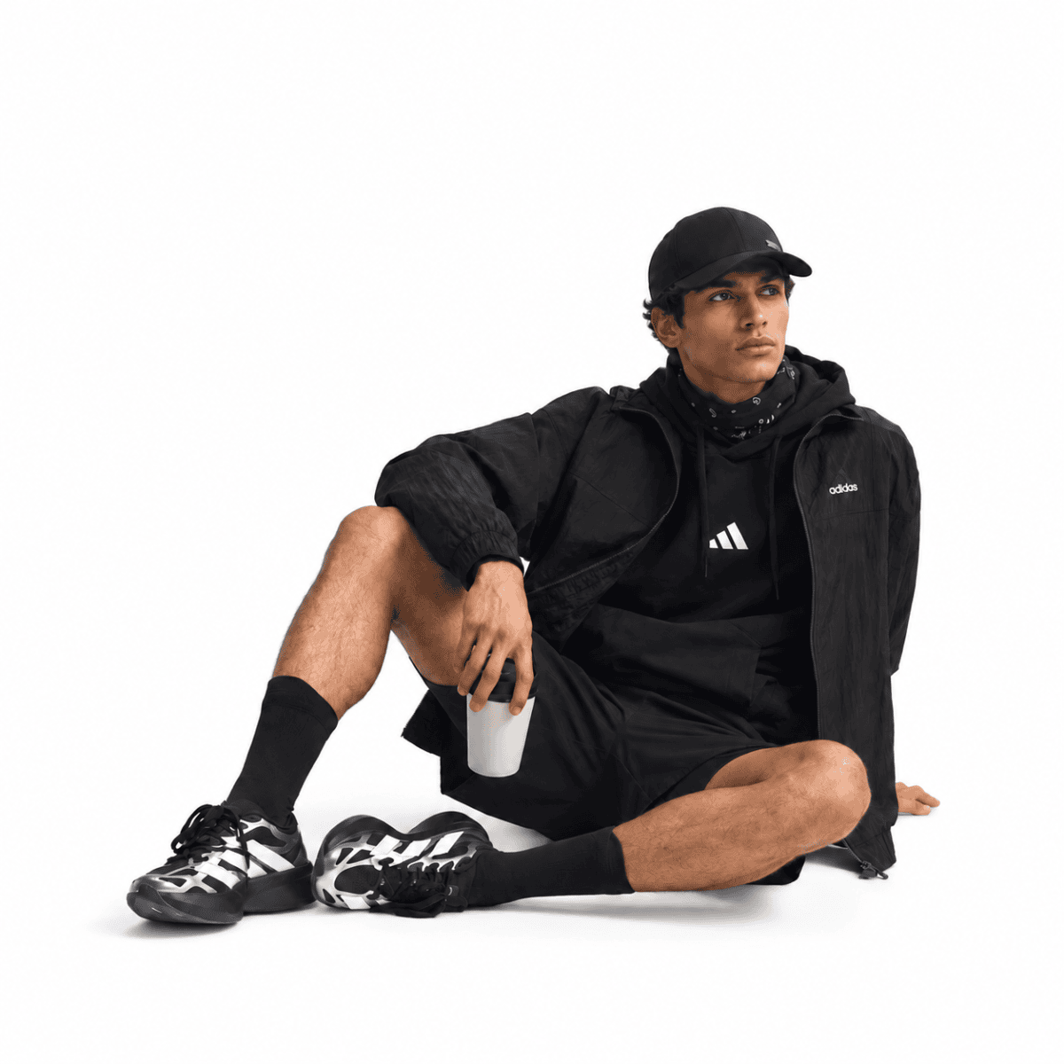 Man in black sportswear sitting with a drink, black cap, and sneakers.