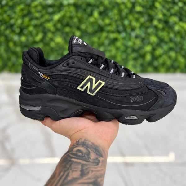 New Balance 1000 triple black sneaker held in hand showing the CORDURA tag and suede panels